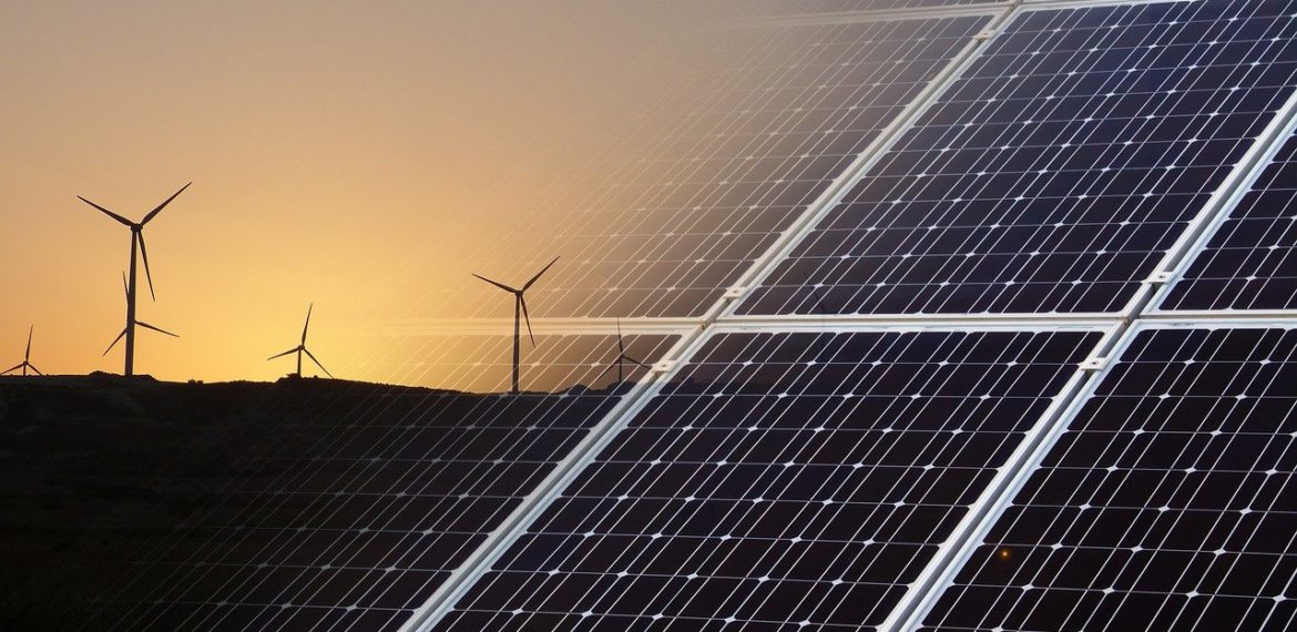 Panneaux solaires au premier plan et éoliennes sur une colline au coucher du soleil, illustrant les énergies renouvelables.